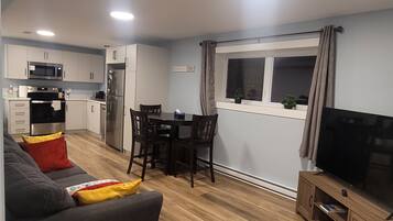 Una cocina moderna con armarios blancos, microondas integrado y refrigerador de acero inoxidable. Cuenta con un comedor con mesa y sillas, y un televisor sobre un mueble de madera. El espacio tiene piso de madera y un sofá gris con cojines rojos y amarillos.