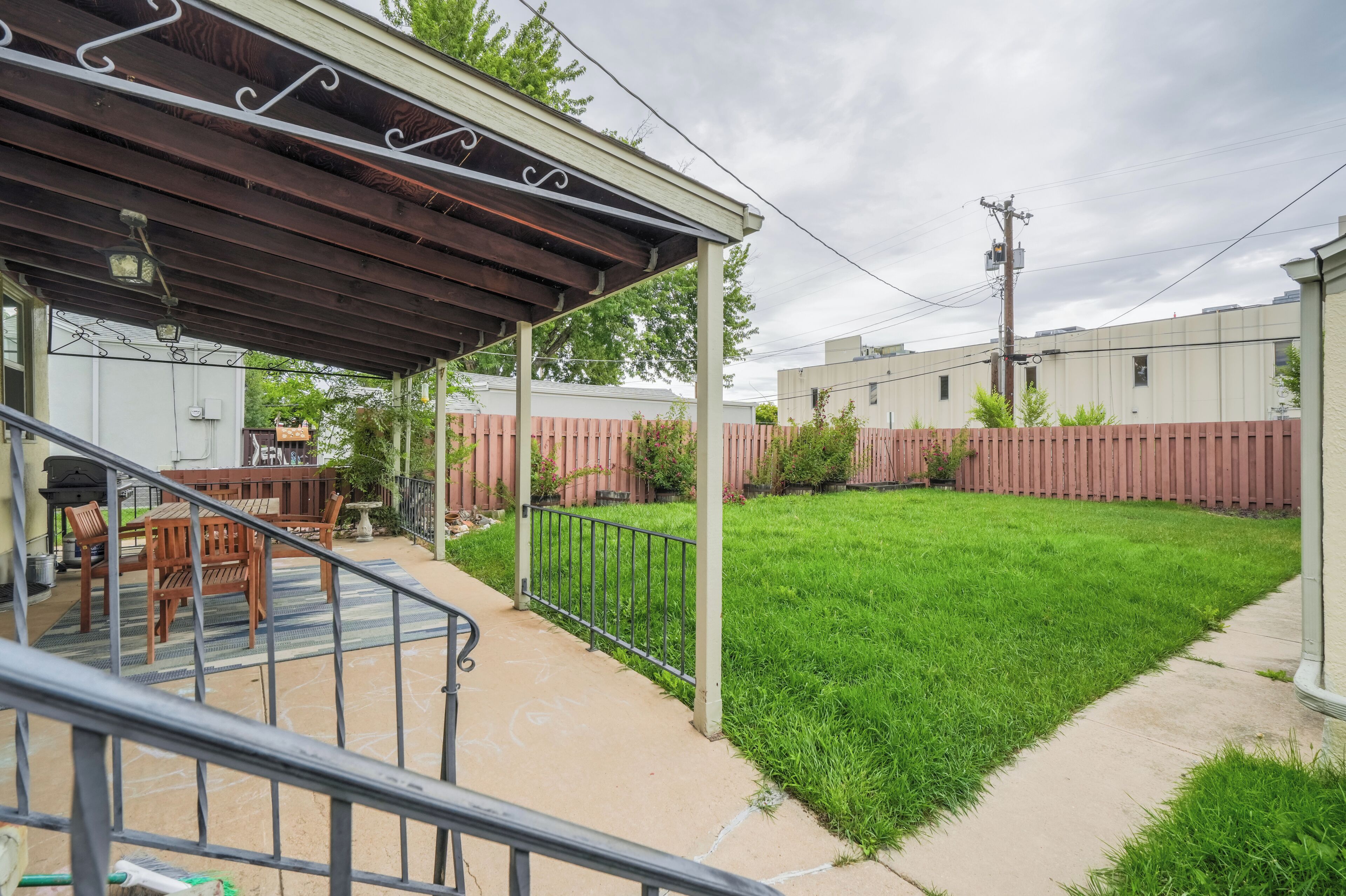 A covered patio area with a wooden table and chairs, a concrete pathway, and a fenced backyard.