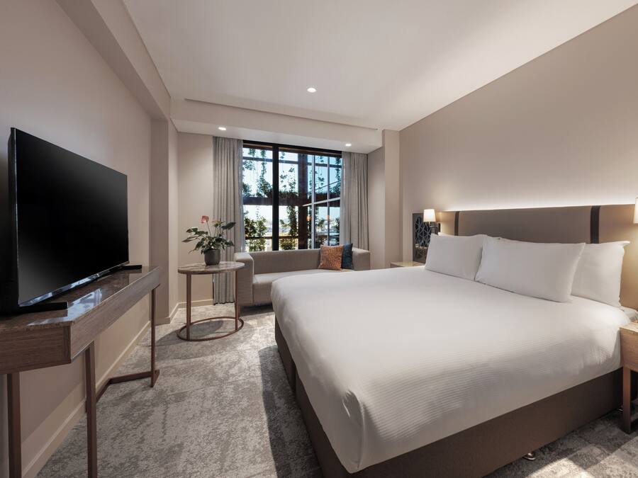 Una habitación de hotel moderna con una cama grande, un televisor de pantalla plana, una mesita, una lámpara y un ventanal con cortinas.