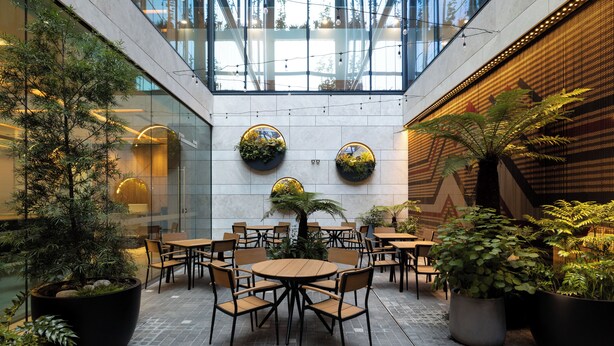 A modern indoor courtyard with round tables and chairs, surrounded by large potted plants and a glass ceiling allowing natural light.