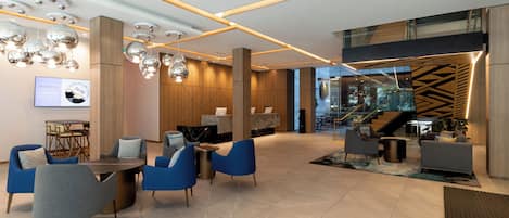 A modern lobby with a reception desk, seating area with blue chairs, and a staircase leading to an upper level.