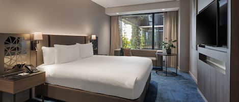 A hotel room with a large bed, a desk with a telephone, a TV, and a view of greenery outside.