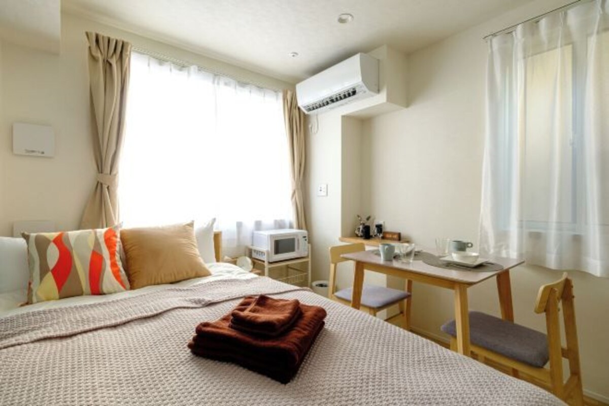 A hotel room with a bed, a desk, a chair, a table, and an air conditioning unit.