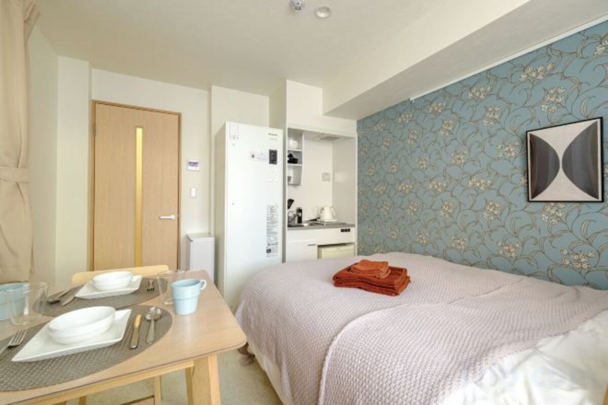 A hotel room with a bed, a desk with a chair, a table with plates and cutlery, a wardrobe, and a bathroom.