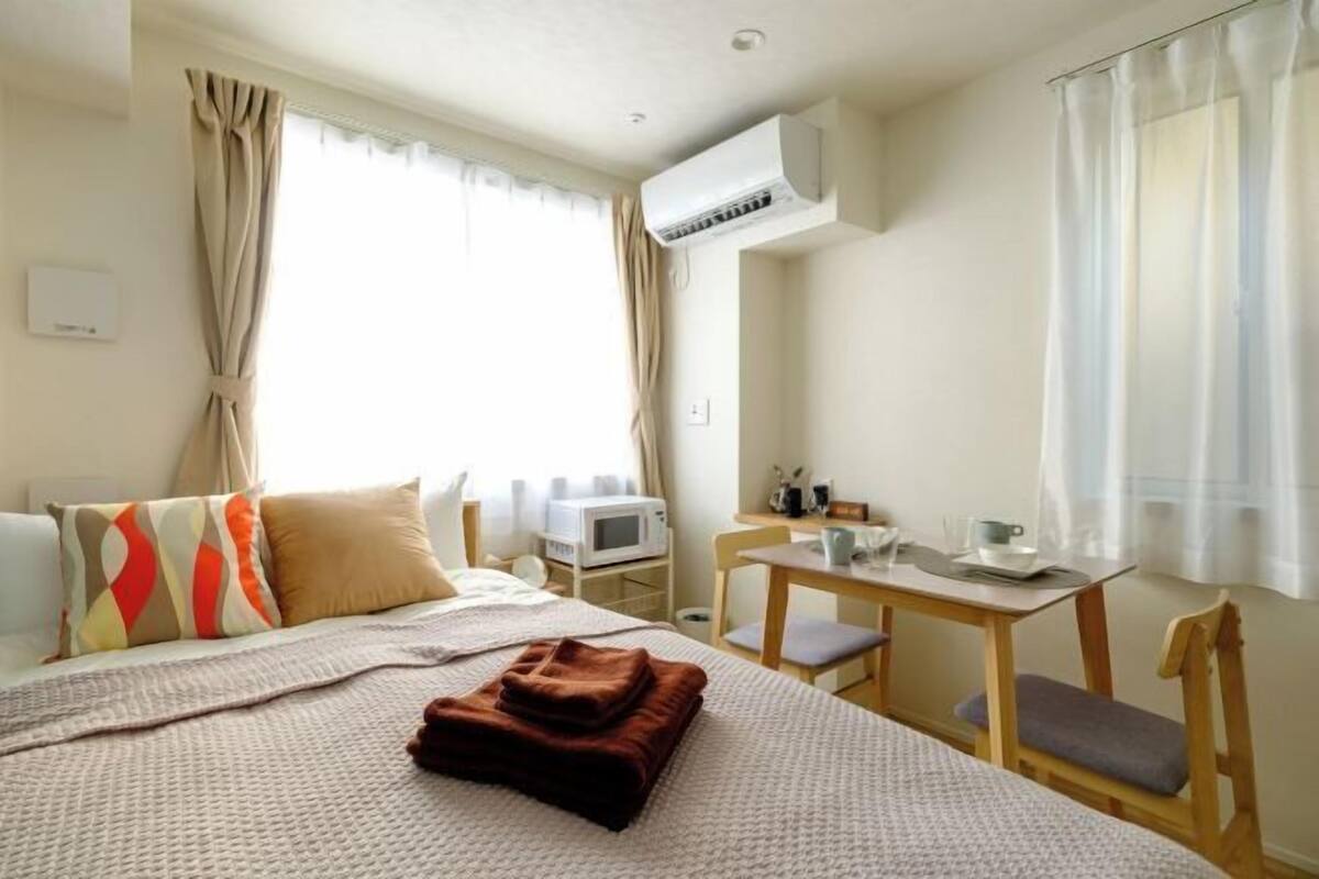 A hotel room with a bed, a desk, a chair, a table, and an air conditioning unit.