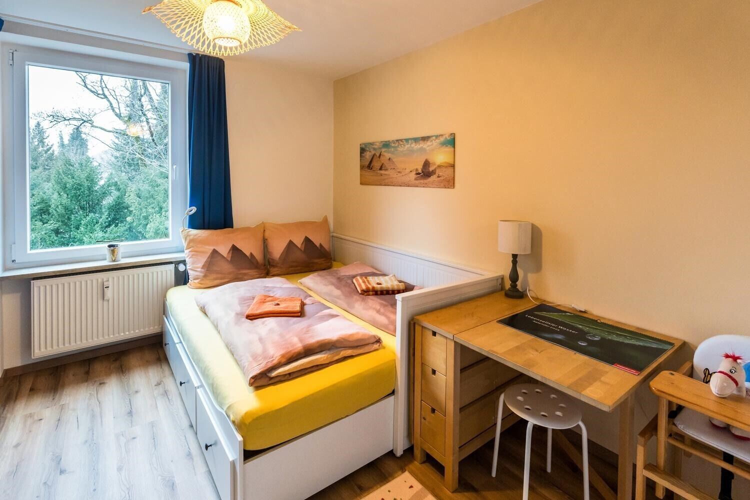 A bedroom with a bed, a desk, a chair, a lamp, a radiator, and a window with a view of trees.