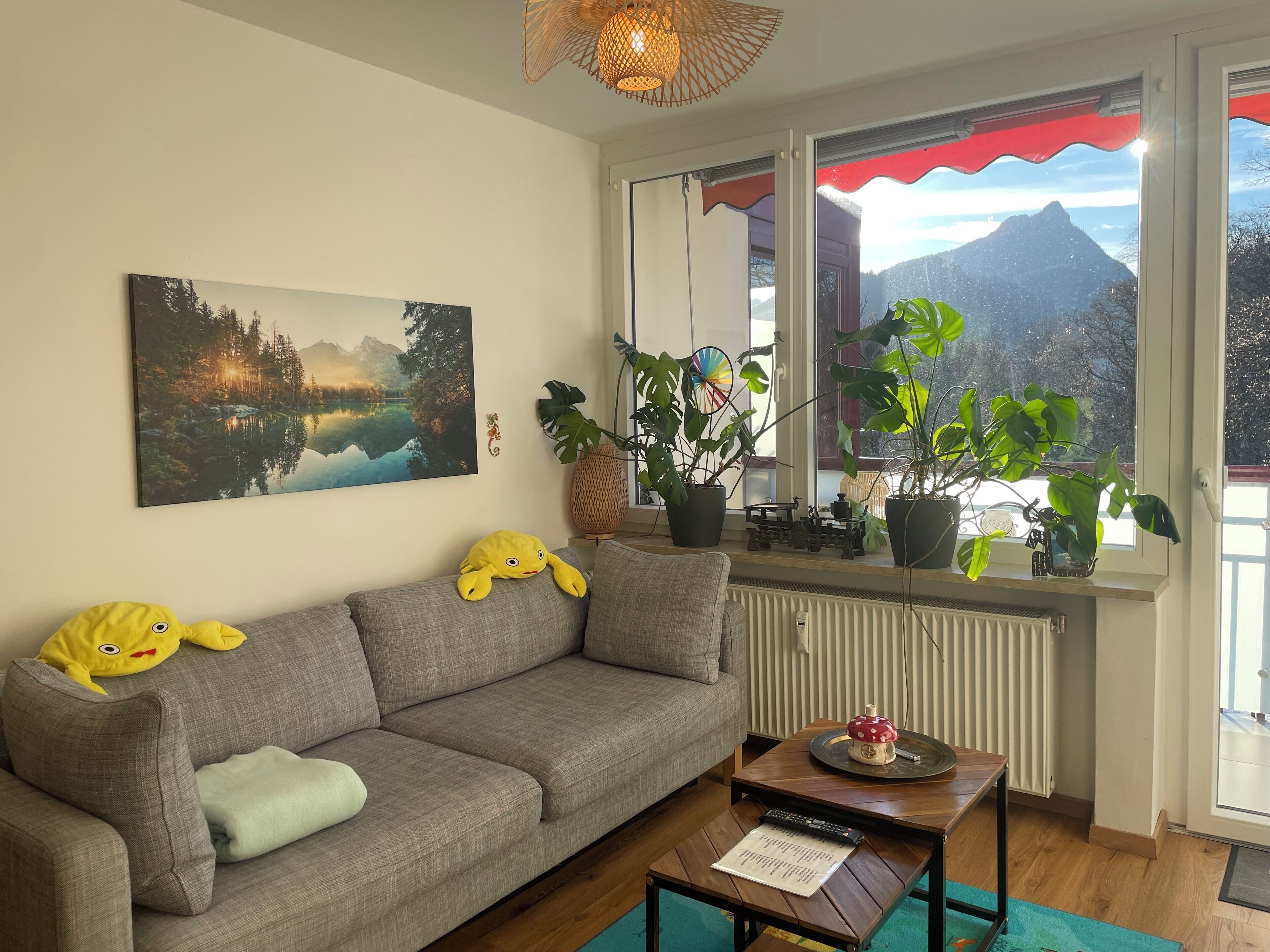 Sala de estar com sofá cinza, brinquedos de pelúcia amarelos, pintura de um lago, plantas grandes em vasos e vista para as montanhas pela janela.