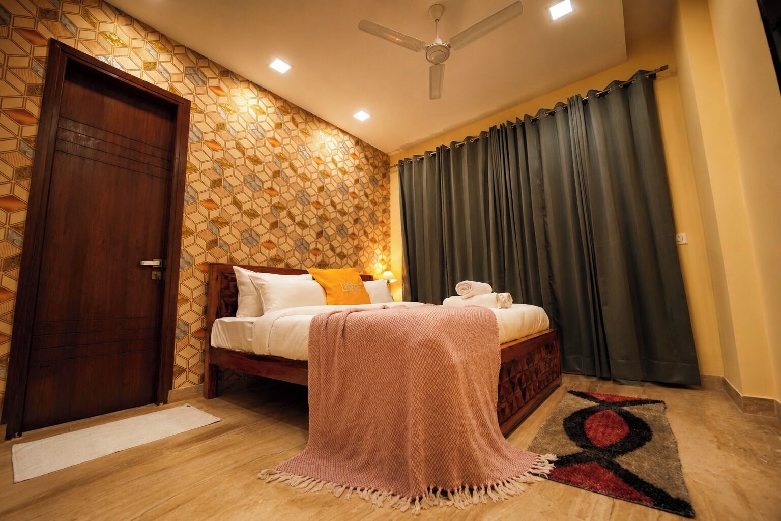 Une chambre avec une porte en bois, un mur à motifs, un lit avec des oreillers, une table de chevet, un ventilateur de plafond et un rideau foncé.