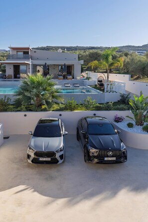 A modern house with a swimming pool, two cars parked in front, and a view of a hilly landscape.
