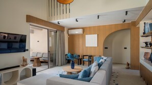En moderne stue med sofa, sofabord og fjernsyn. Der er en trappe, der fører til et andet plan, og en trævæg med en aircondition-enhed.