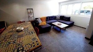 En stue med et mønstret bord, en mørk sofa og et vindue med udsigt til parkerede biler.