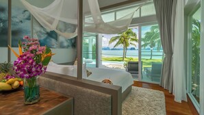 A bedroom with a large bed, a view of the ocean, a tropical-themed painting, and a vase of flowers on a wooden table. - Coastal Escape Koh Yao Noi (Ko Yao)