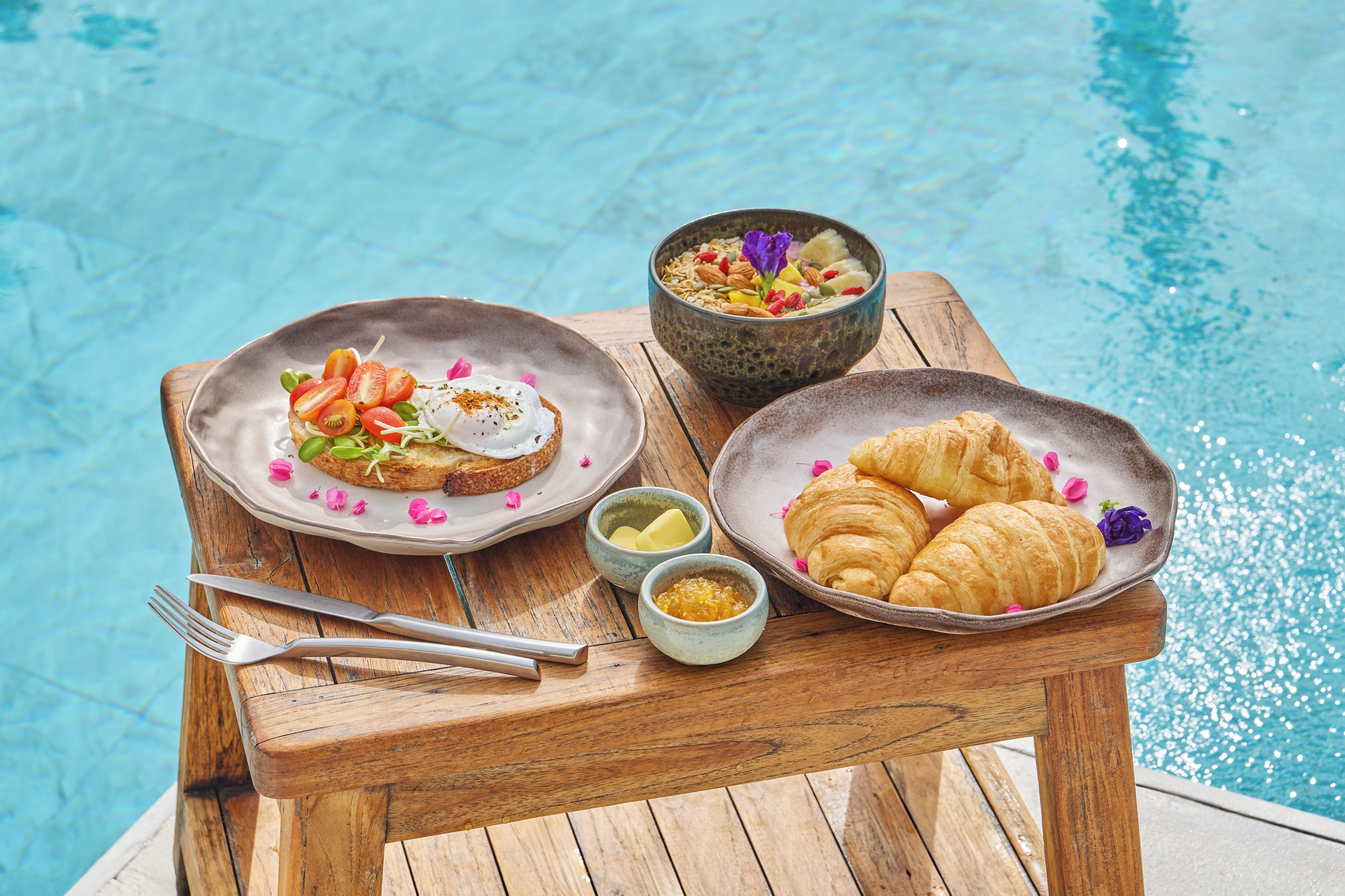 Une table en bois, dressée avec des aliments pour le petit-déjeuner, se trouve au bord de la piscine.