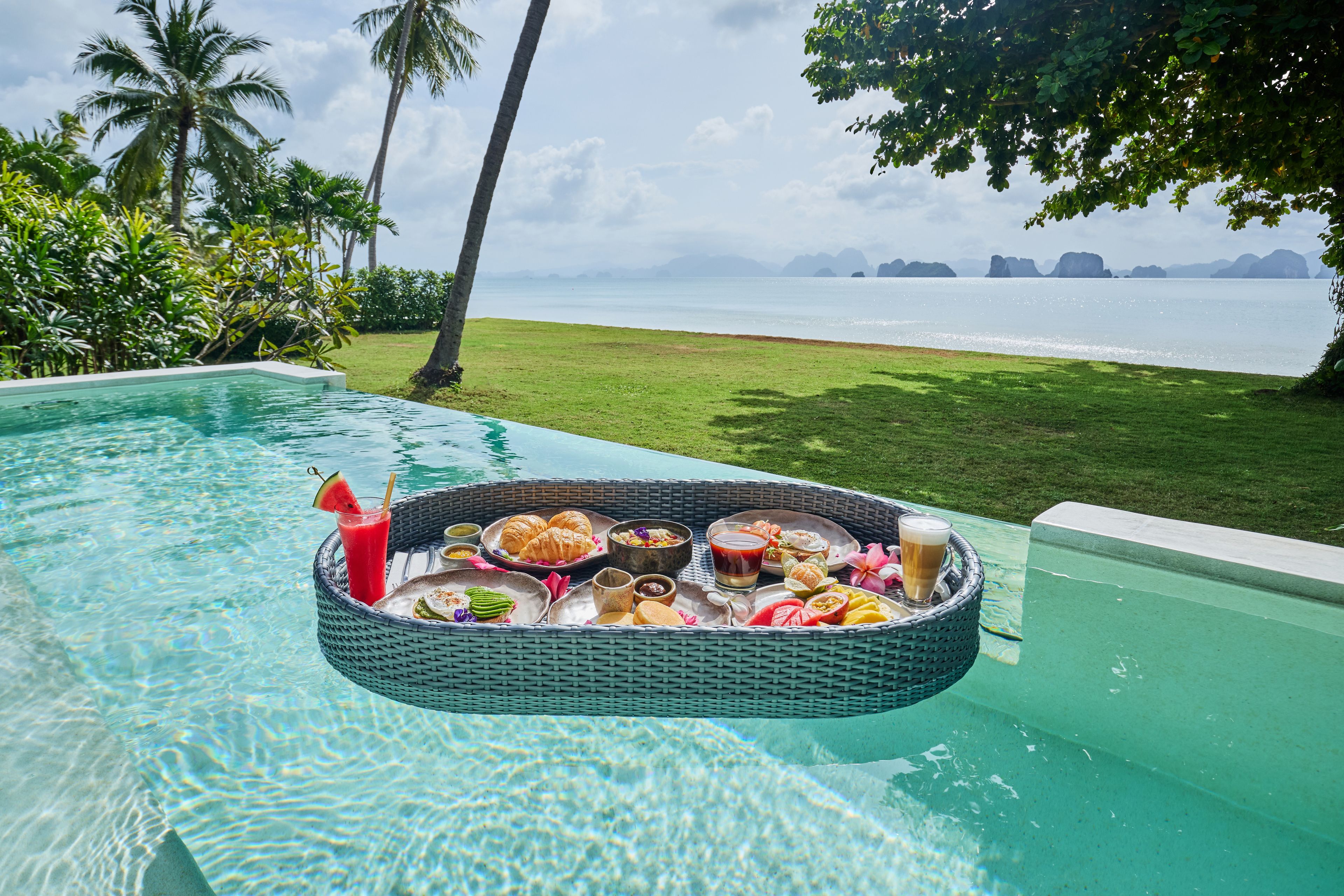 A floating dining table with a variety of food and drinks, overlooking a pool and a view of the sea with palm trees and distant islands.