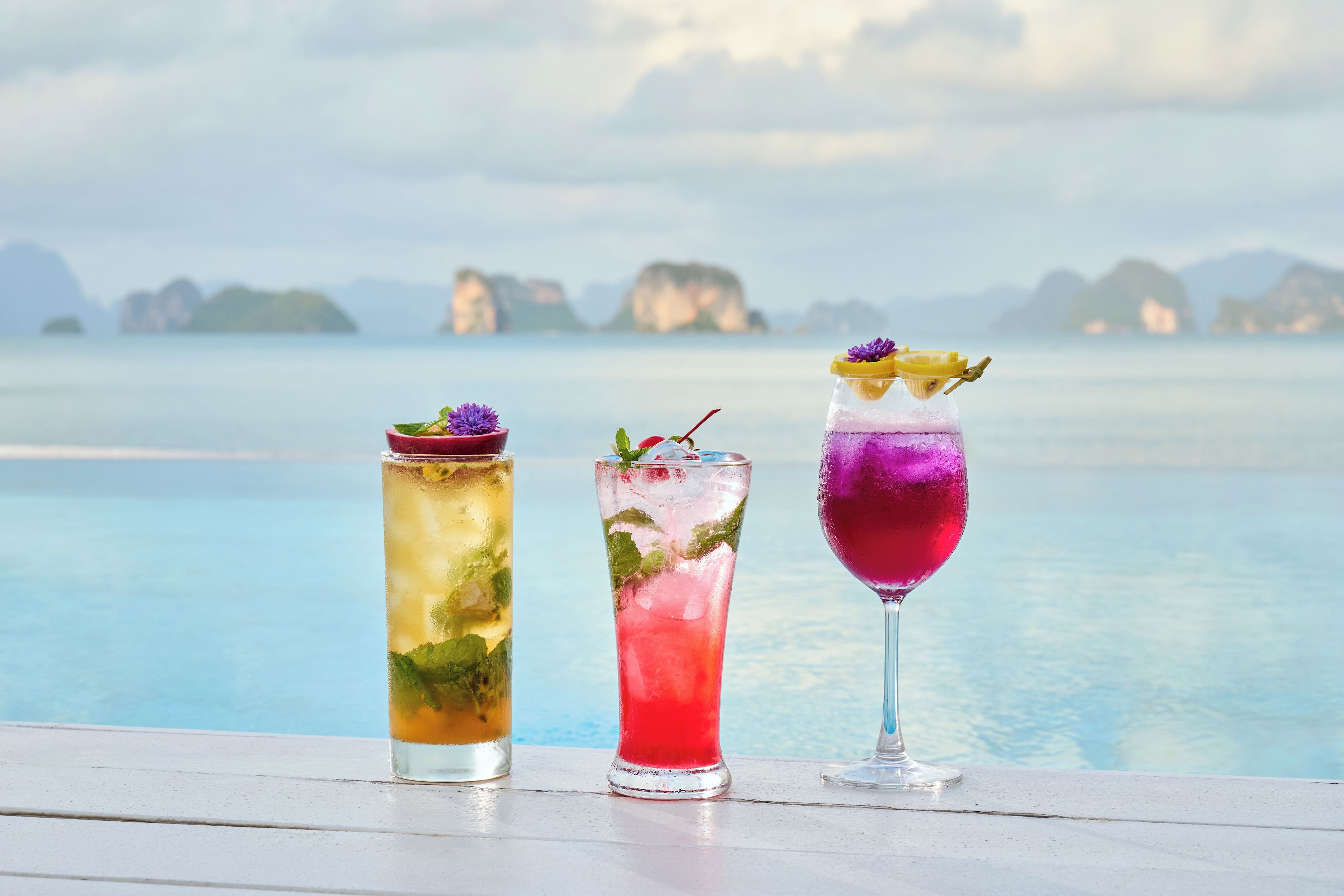 Three cocktails on a white surface with a view of a calm sea and distant islands.