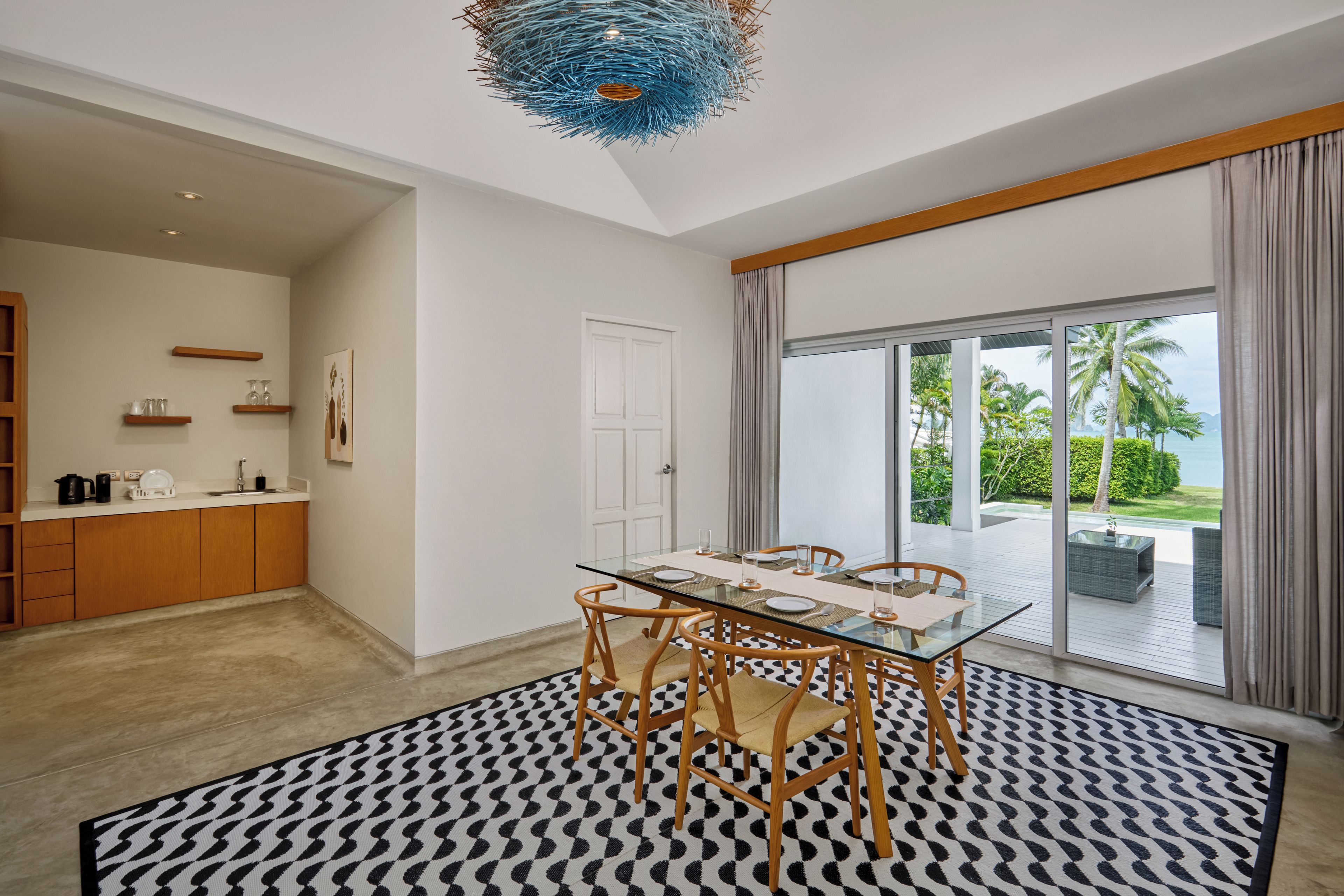 A modern dining area with a glass table and wooden chairs, a kitchen with wooden cabinets, and a large sliding glass door leading to a balcony with a view of palm trees and the sea.