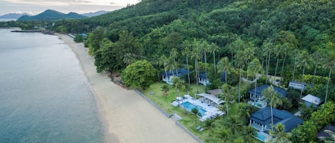 An aerial view of a coastal resort with a beachfront, lush greenery, and multiple villas with swimming pools.