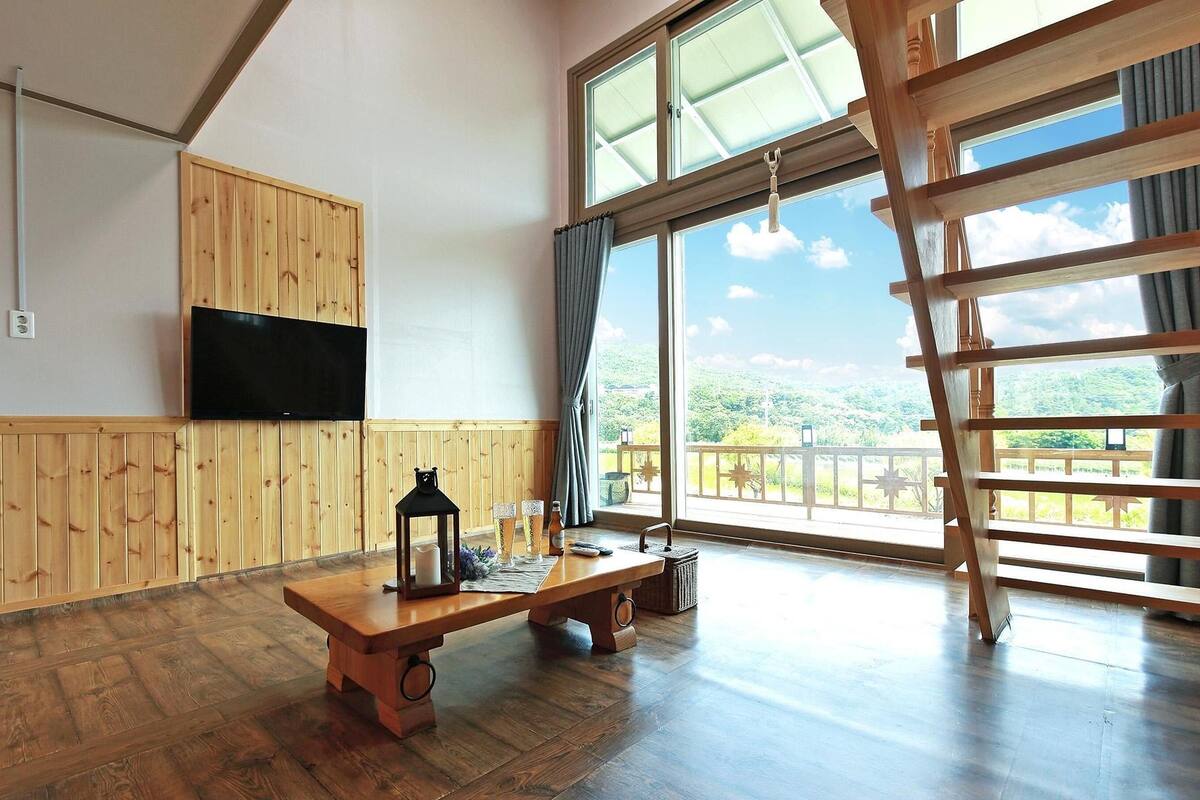 A room with wooden panel walls, a flat-screen TV mounted on the wall, a wooden bench with a lantern, and a large window with a view of greenery.