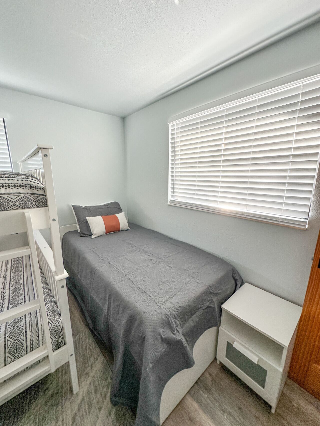 A small, neatly arranged bedroom with a single bed, a nightstand, a bedside table, and a crib.