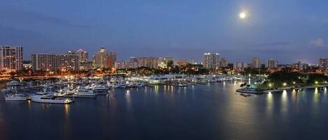 A marina with boats docked, a city skyline in the background, and a moon in the sky.