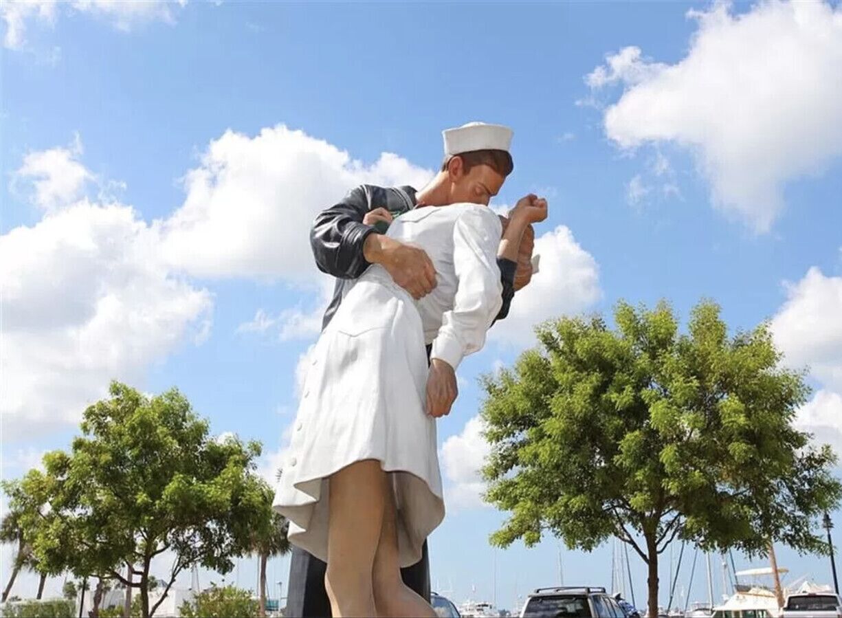 A person in a white uniform lifting another person in a white dress outdoors with trees and boats in the background.