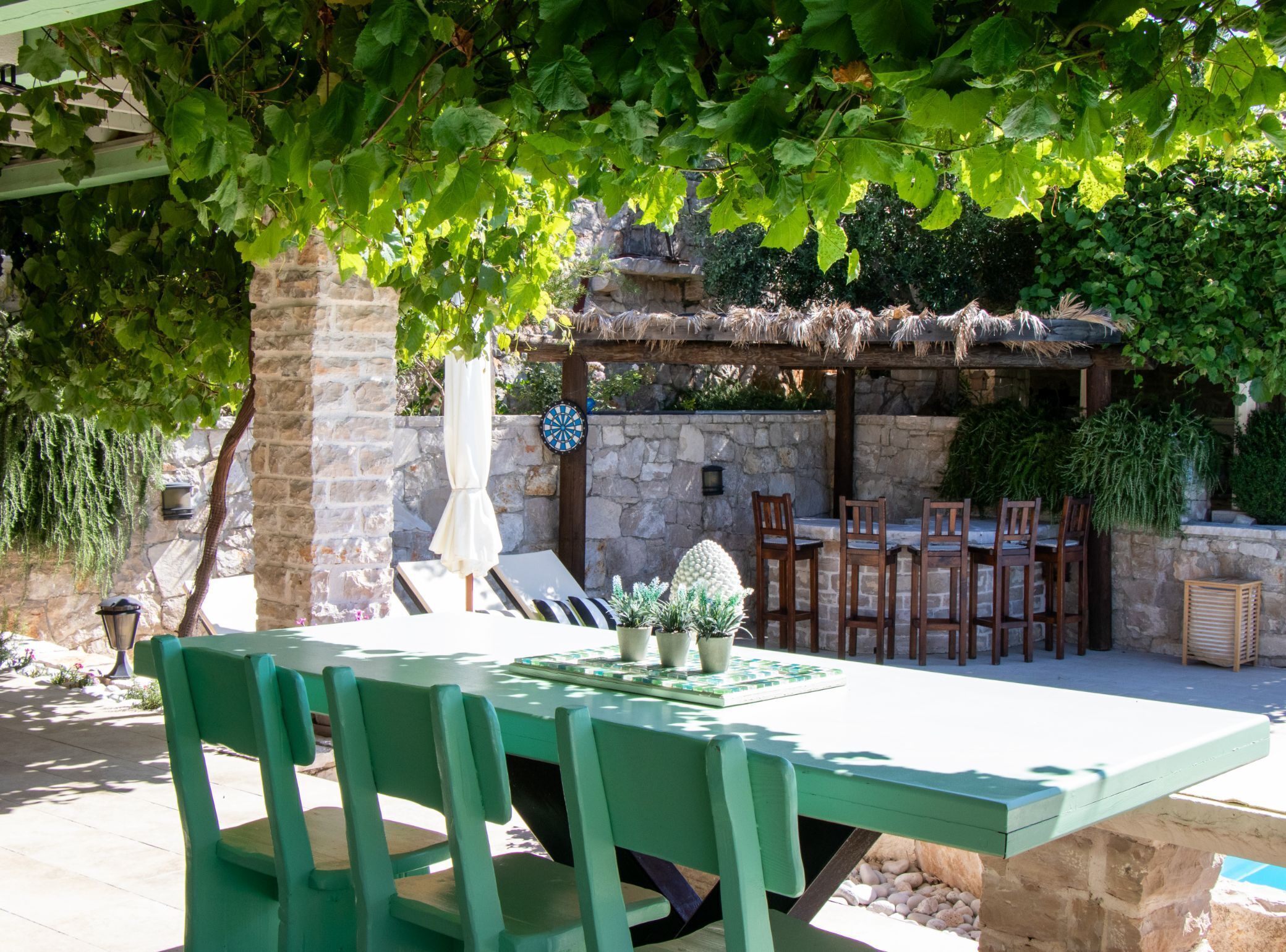 A green wooden table and chairs under a tree, with a stone wall and a thatched roof structure in the background.