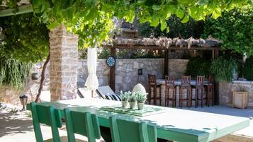 A green wooden table and chairs under a tree, with a stone wall and a thatched roof structure in the background.
