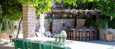 A green wooden table and chairs under a tree, with a stone wall and a thatched roof structure in the background.