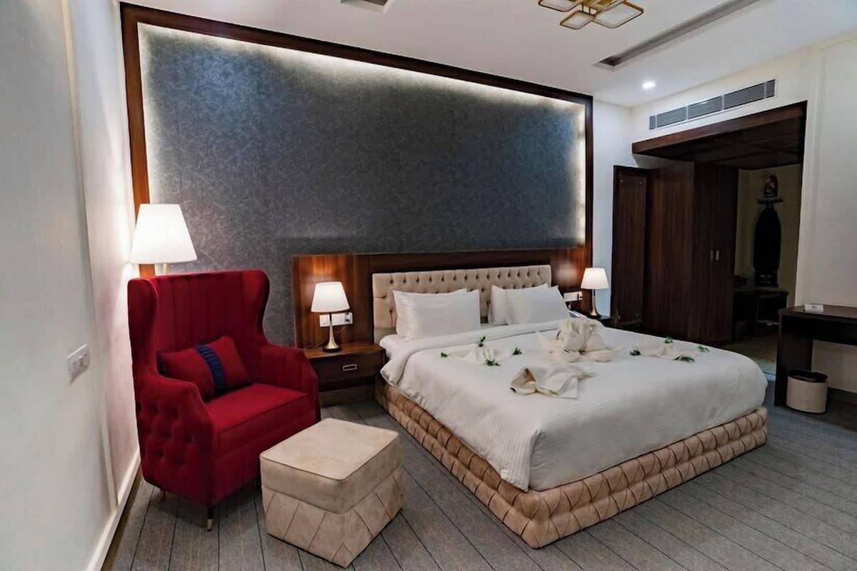 A hotel room with a large bed, a red armchair, a small ottoman, and a wooden headboard.