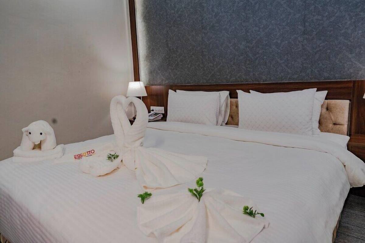 A hotel room with a neatly made bed, a lamp on the nightstand, and a towel rack with towels.