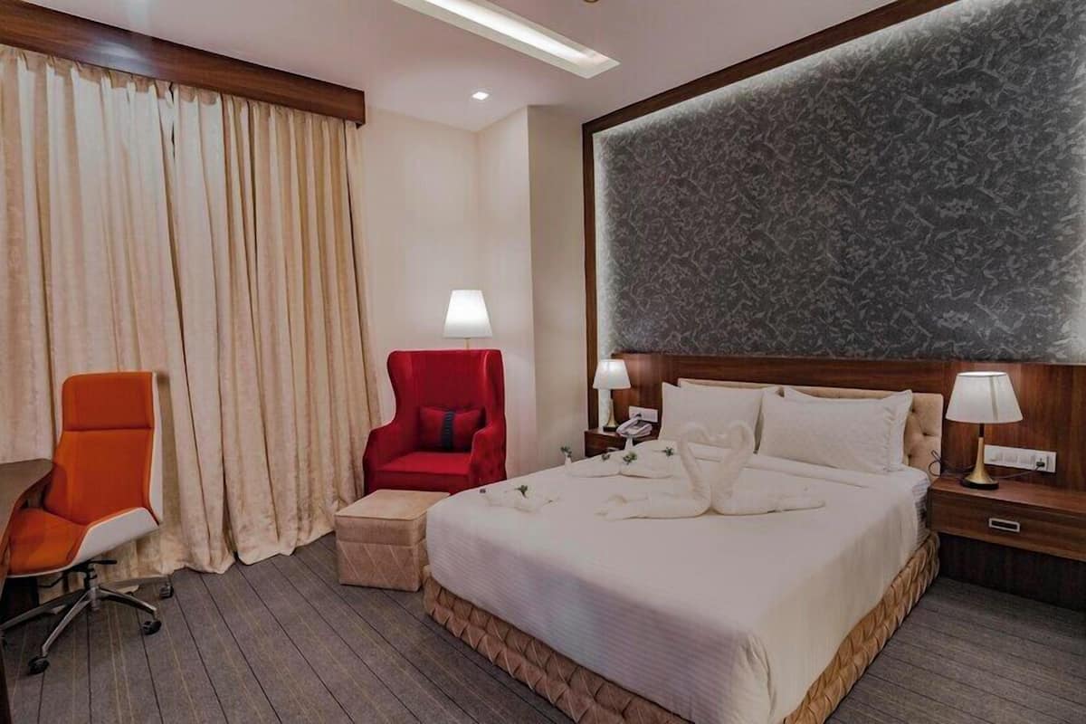 A hotel room with a large bed, a red armchair, a small ottoman, a desk with a chair, and a large decorative wall panel.