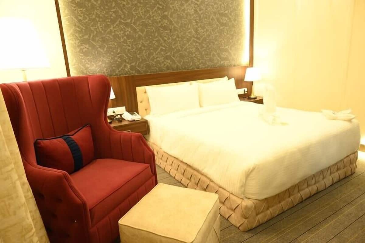 A hotel room with a large bed, a red armchair, a small bedside table, and a nightstand with a lamp.