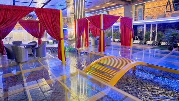 A luxurious indoor pool area with red drapes, a glass floor, and a wooden bench. - Essotto Recreation Hub (Bengaluru)