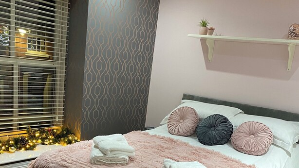 A modern bedroom with a large bed, a decorative wall, a window with blinds, and a small shelf. - Snaith Central Apartment (Goole)