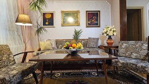 A living room with a sofa, two armchairs, a coffee table with a fruit bowl, a lamp, and framed artwork on the wall. - Spacious, stylish and cozy apartment in the very center of Sofia (София)