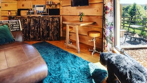 A cozy cabin interior with a wooden ceiling, a television mounted on the wall, a bar area with stools, and a large window offering a view of the outdoors. - Sweet Donside Cabins (Strathdon)