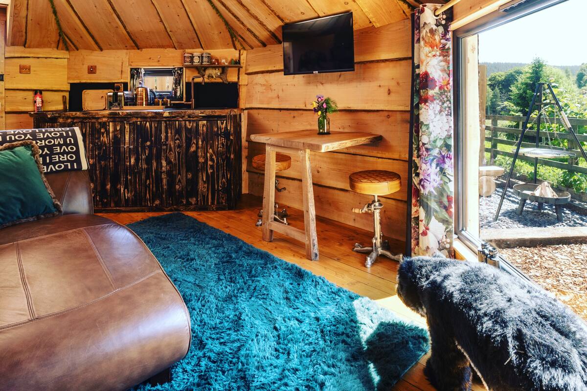 A cozy cabin interior with a wooden ceiling, a television mounted on the wall, a bar area with stools, and a large window offering a view of the outdoors.