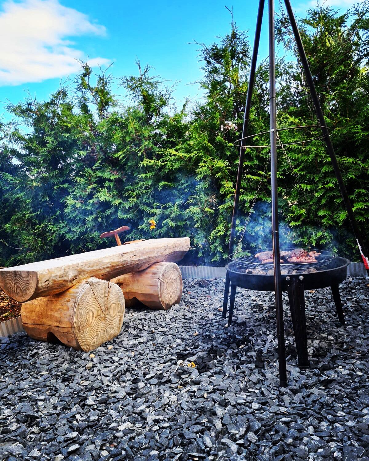 Ein Grillbereich im Freien mit Holz und Feuerstelle, umgeben von Kies und Grünpflanzen.
