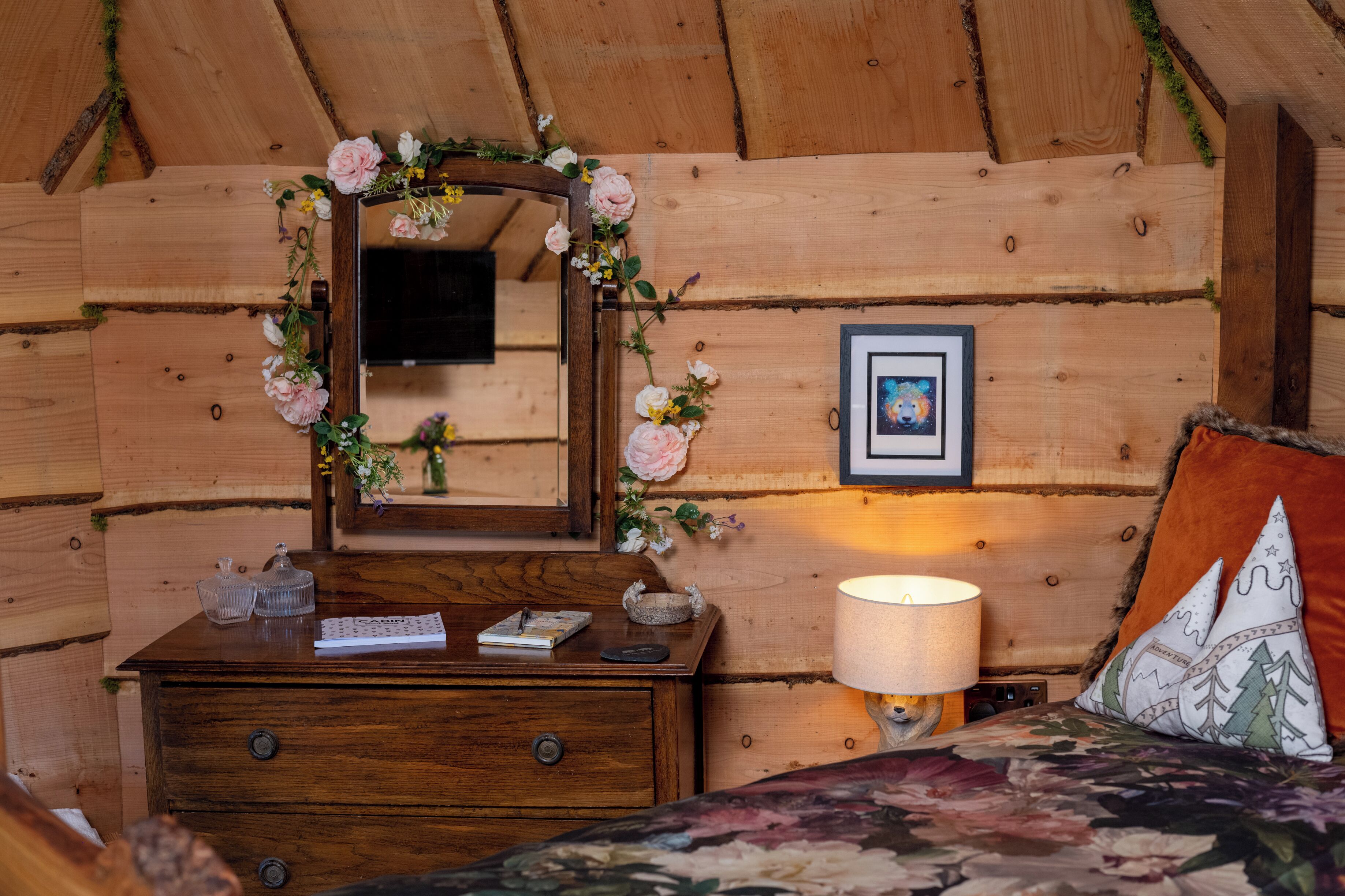 Habitación de cabaña de madera con cama, cómoda, espejo con adornos florales, un cuadro enmarco y una lámpara.