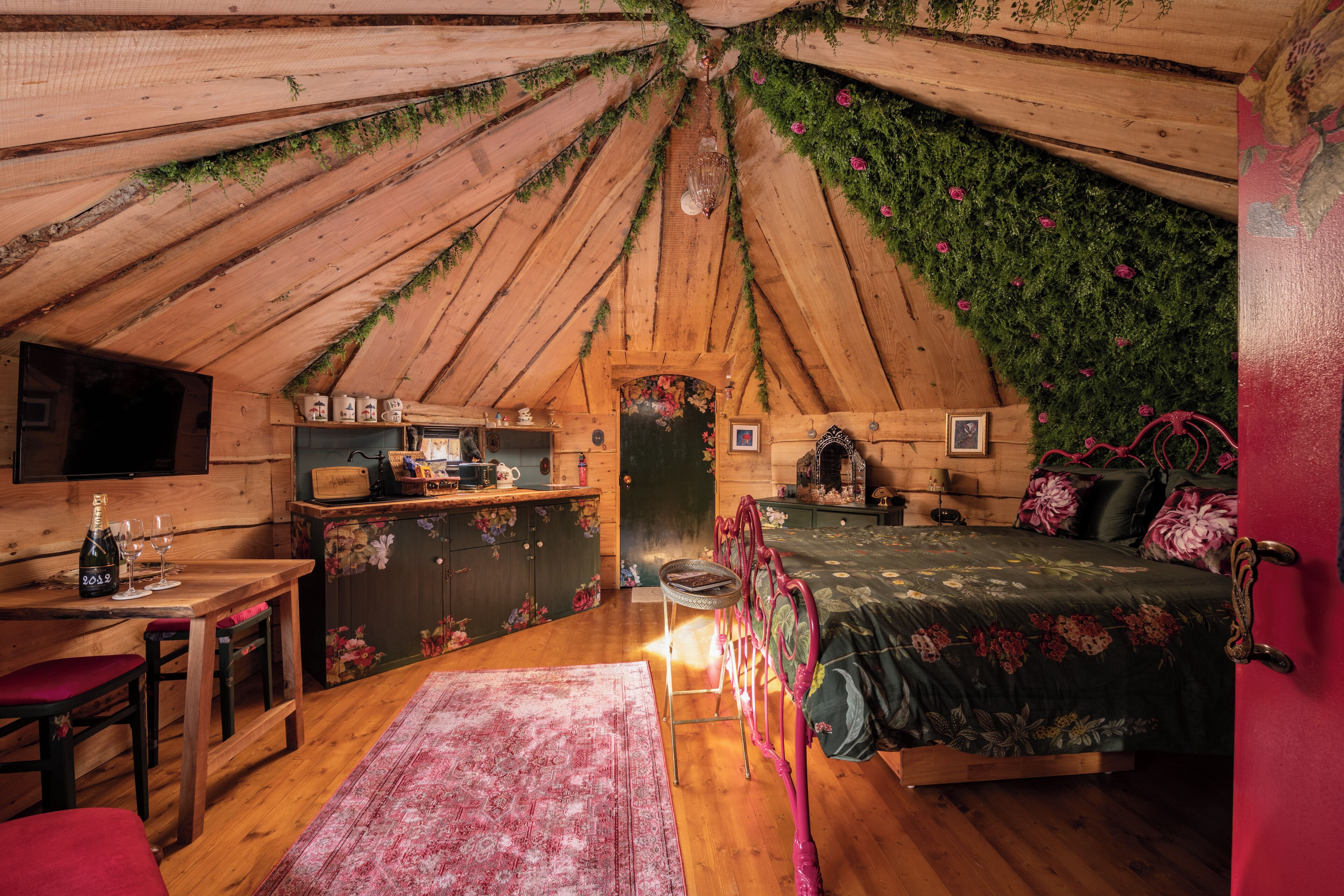 Un dormitorio acogedor en un ático con techo inclinado de madera, una cama con ropa de cama floral, un escritorio de madera con una botella y vasos, y una mesita.