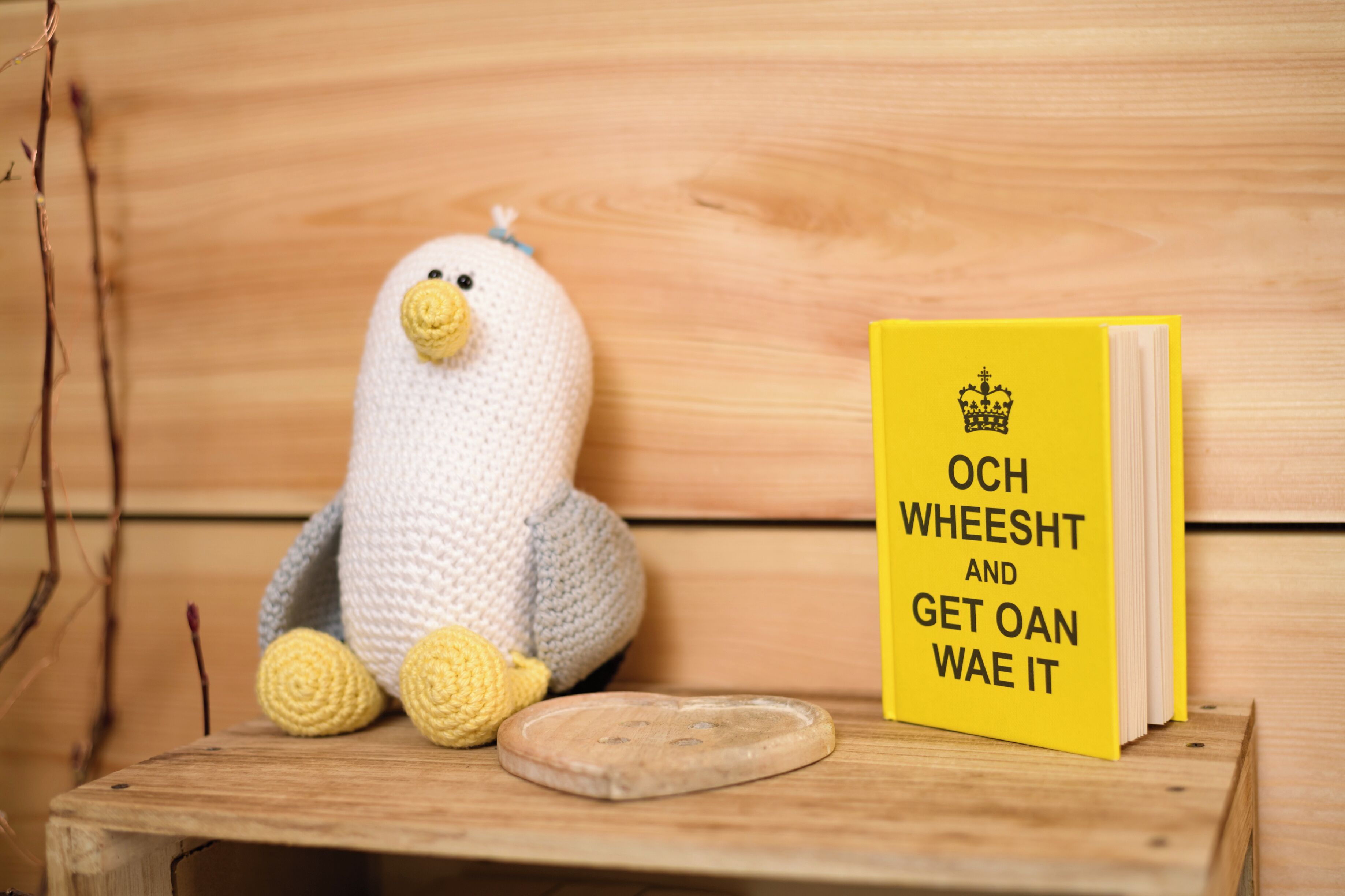 Een houten bank met een gehaakte pinguïn en een geel boek getiteld 'Och WHEESHT AND GET OAN WAE IT'.