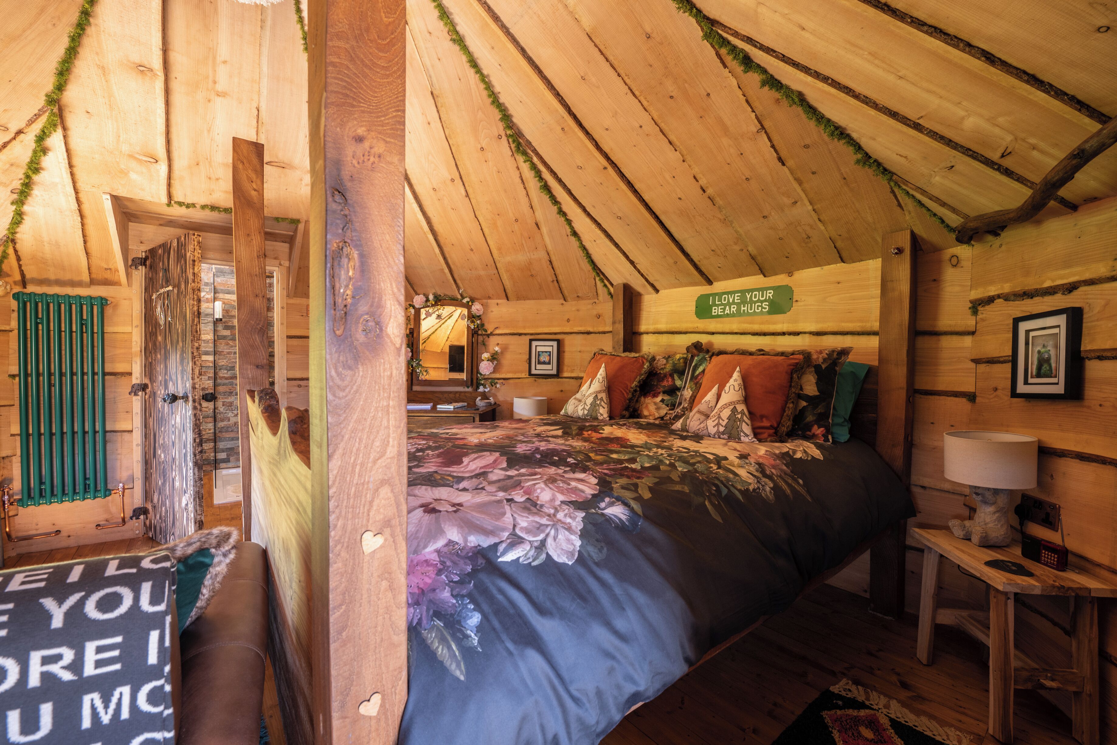 A cozy attic bedroom with a wooden ceiling, a bed with floral bedding, and a sign that says "I love your bear hugs".