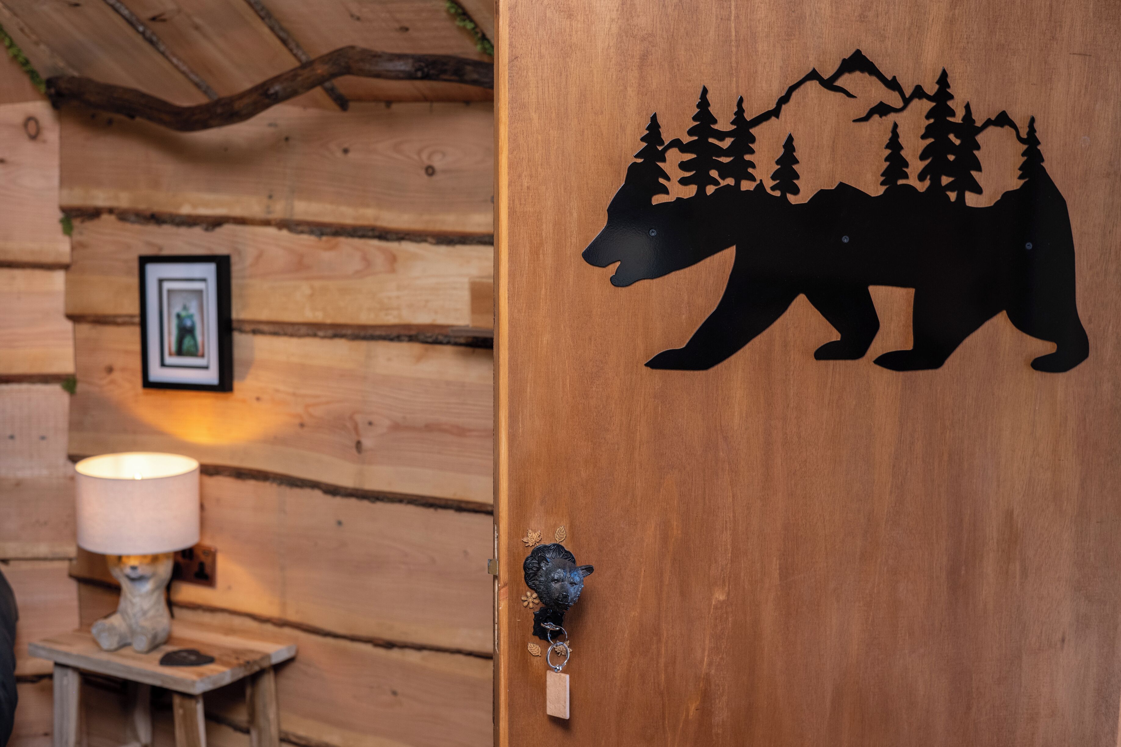 Uma porta de madeira com silhueta de urso e paisagem de montanha, um quadro emoldurado na parede e um abajur sobre uma mesa pequena.