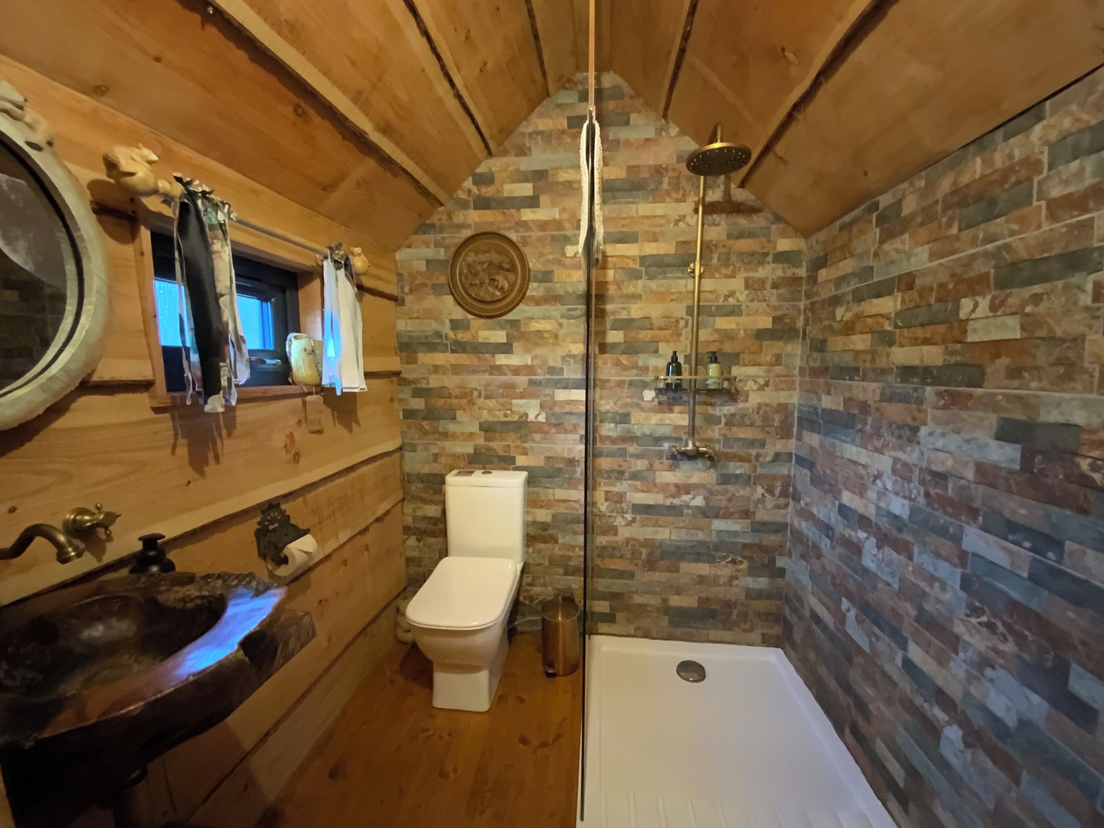 A wooden cabin bathroom with a stone wall, a wooden sink, a toilet, and a shower.
