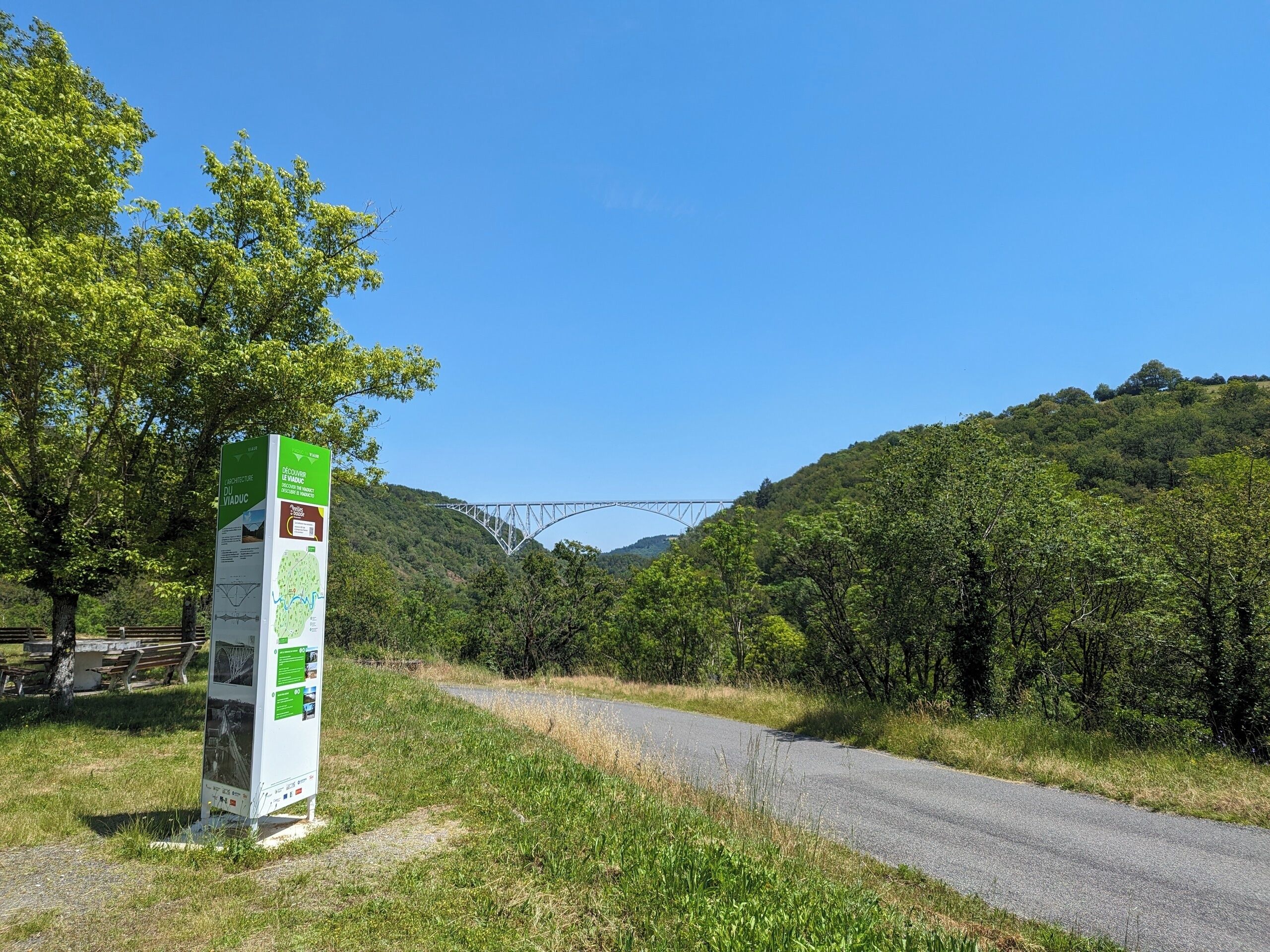 A signpost with information on a rural road, surrounded by greenery and a bridge in the background.