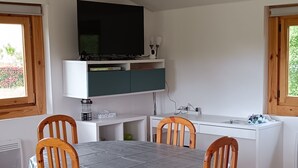 Spiseområde med bord, stole, et fjernsyn over bordet og et køkken i baggrunden.
