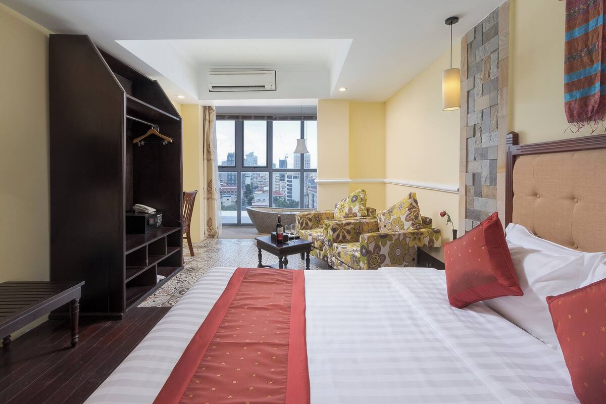 A hotel room with a large bed, a wardrobe, a seating area with chairs, a small table, and a view of the city through large windows.