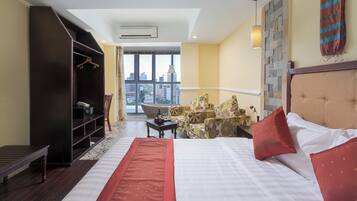 Habitación de hotel con una cama grande, un armario, una zona de estar con sillas, una mesita y vistas a la ciudad a través de amplios ventanales.