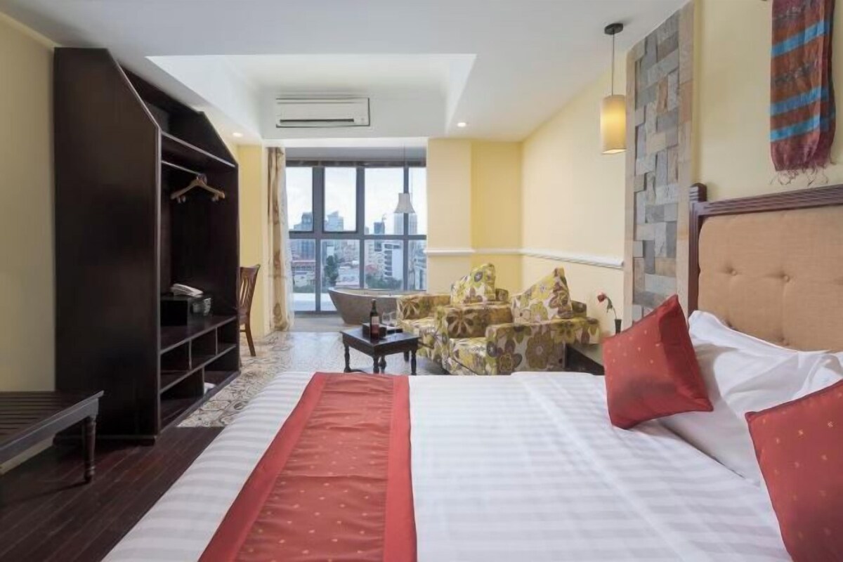 A hotel room with a large bed, a seating area with chairs, a small table, and a view of the city through large windows.
