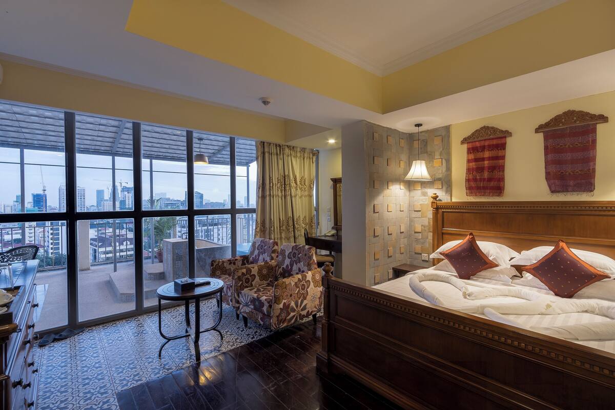 A hotel room with a large window, a bed with decorative pillows, a wooden headboard, and a small round table with a chair.